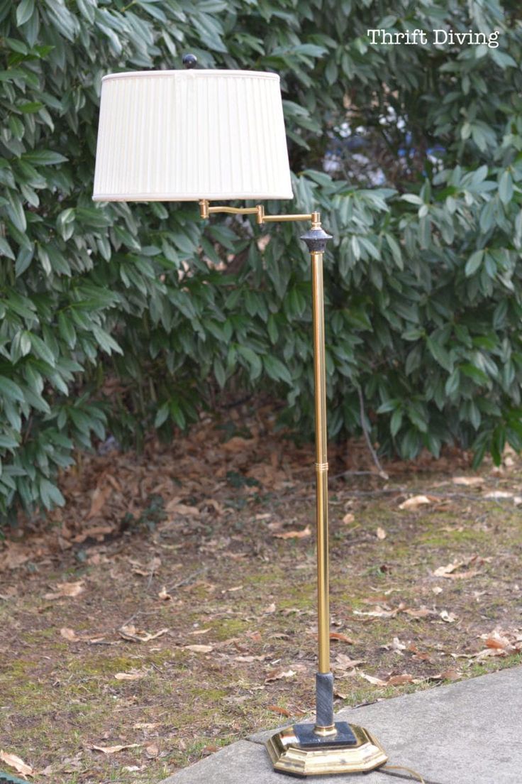 a floor lamp with a white shade on it in front of some bushes and trees