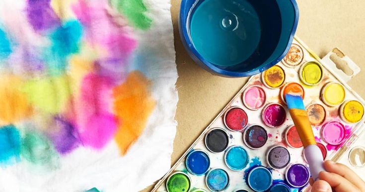 a child's hand holding a paintbrush next to an art project with watercolors