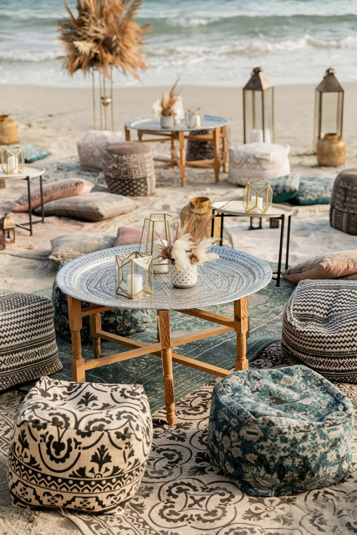 an outdoor seating area on the beach with lots of pillows and stools around it