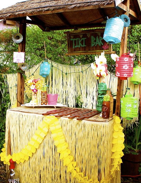 a tiki bar decorated with yellow and blue paper lanterns, grass decorations and flowers