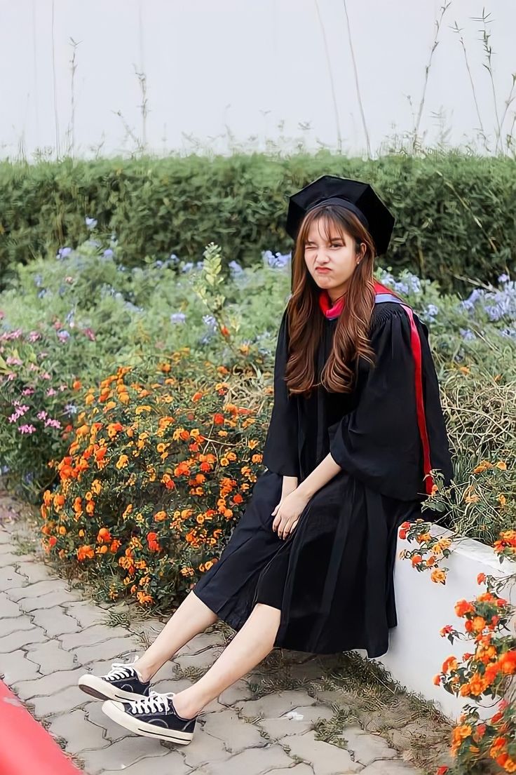 a woman sitting on the ground wearing a black graduation gown and hat with flowers in the background