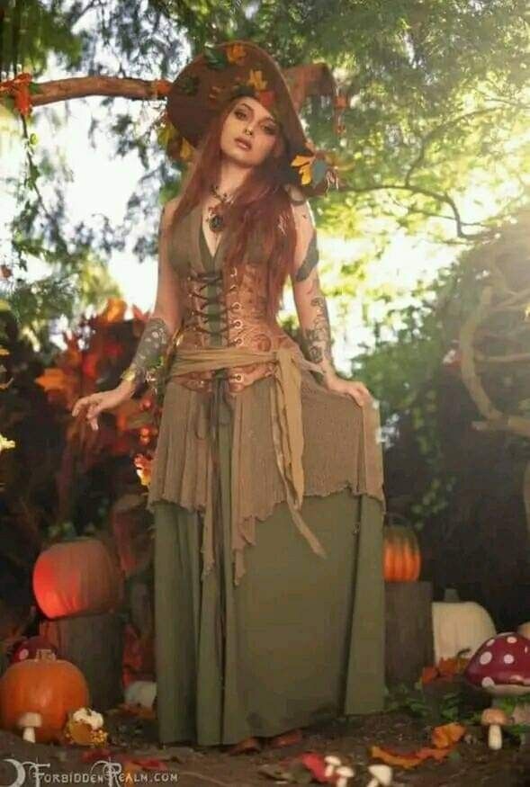 a woman dressed up as a witch standing in front of some pumpkins and trees