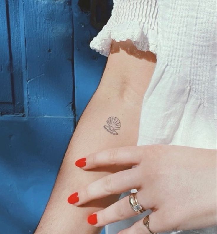 a woman's hand with a small shell tattoo on her left wrist and red nail polish