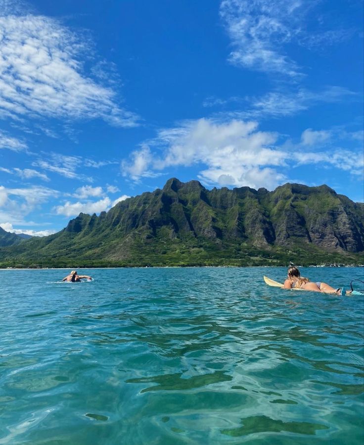 two people on surfboards in the ocean with mountains in the background and blue sky