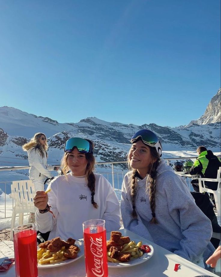 two women sitting at a table with food and drinks in front of snow covered mountains