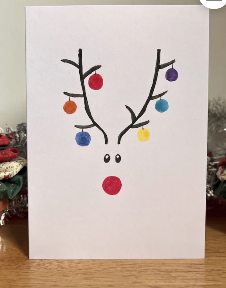 a christmas card with a reindeer's face painted on the front and colorful balls hanging from its antlers