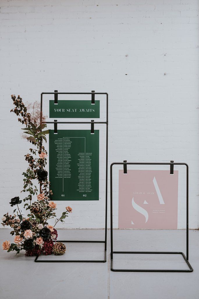 two black metal stands with flowers on them against a white brick wall, one holding a menu and the other displaying an event plan