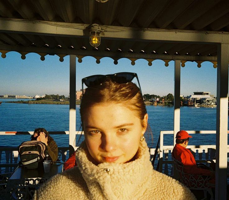 a woman standing in front of a lake with people sitting at tables and looking into the camera