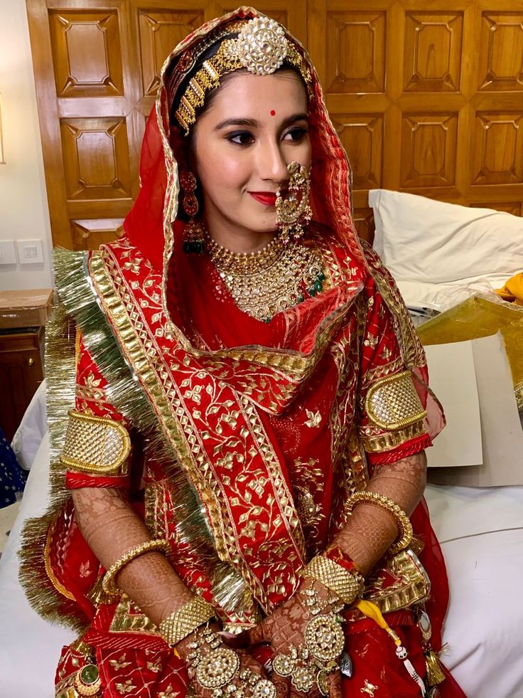 a woman in a red and gold bridal outfit sitting on a bed wearing jewelry