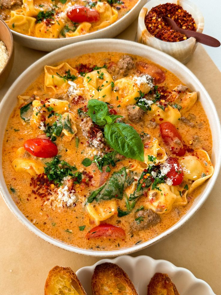 two white bowls filled with pasta and vegetables on top of a table next to bread