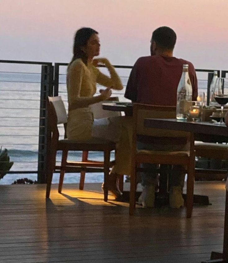 two people sitting at a table with wine glasses in front of them and the ocean behind them