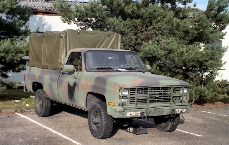 an army truck is parked in a parking lot near some pine trees and a building