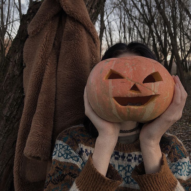 a person with a pumpkin on their head in front of a tree and wearing a jacket