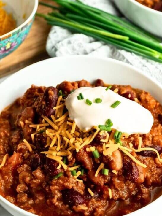 a white bowl filled with chili and cheese on top of a wooden table next to green onions