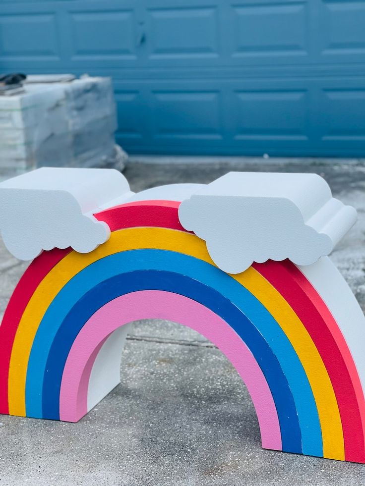 a wooden toy with a rainbow painted on the side and clouds in the middle, sitting next to a blue garage door