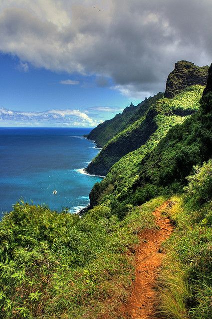 a dirt path going up the side of a mountain next to water and clouds in the sky