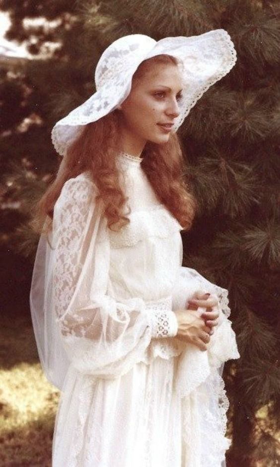 a woman in a white dress and hat standing next to a tree with her hands on her hips