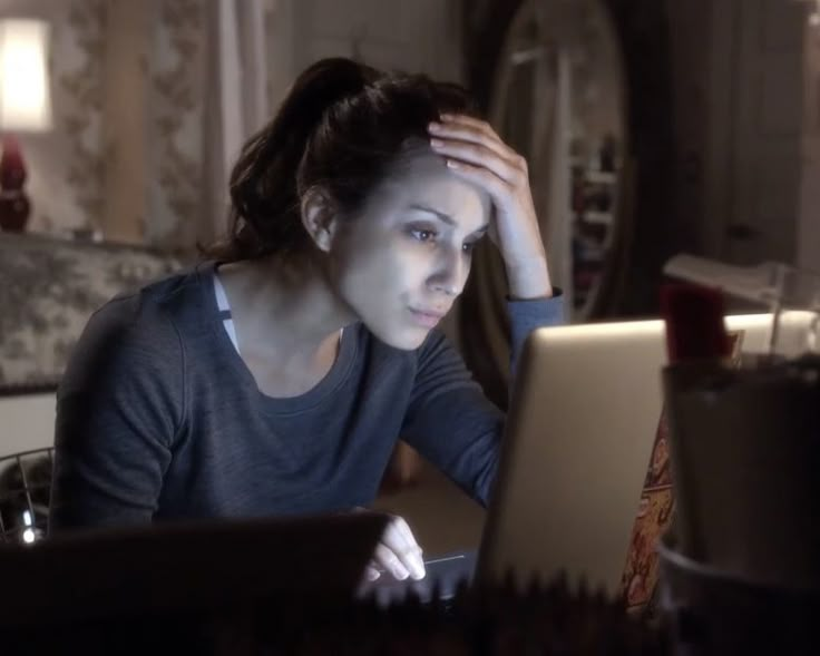 a woman sitting in front of a laptop computer with her hand on her head and looking at the screen