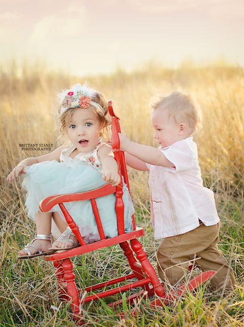 two young children sitting in a red rocking chair on the grass with their hands touching each other