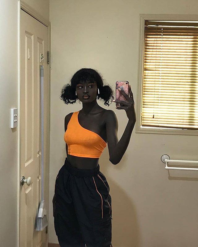 a woman taking a selfie with her cell phone in front of the camera, wearing an orange top and black shorts