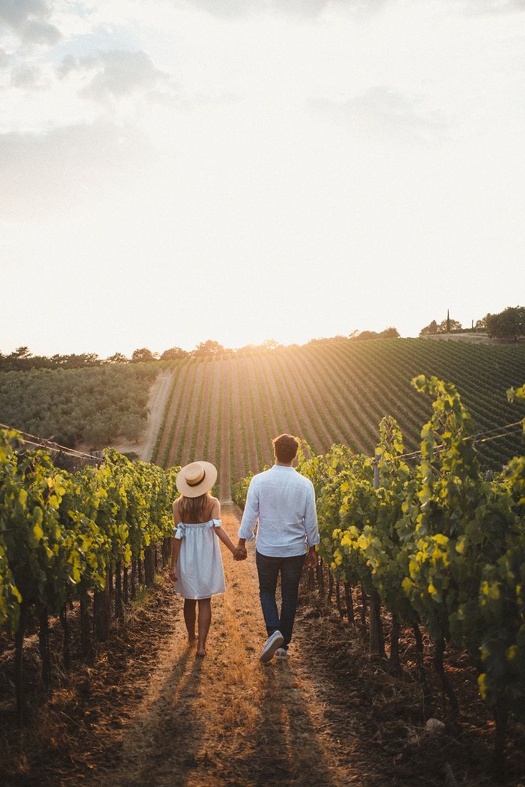 a man and woman walking down a dirt road in the middle of a vineyard field