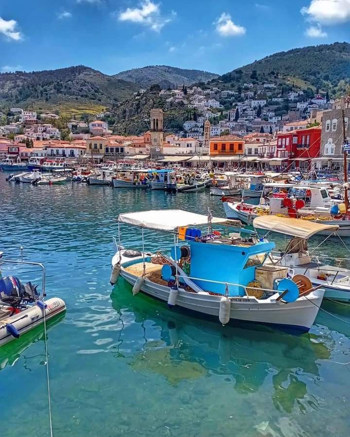 several small boats are docked in the water near some buildings and hills, with blue skies above them