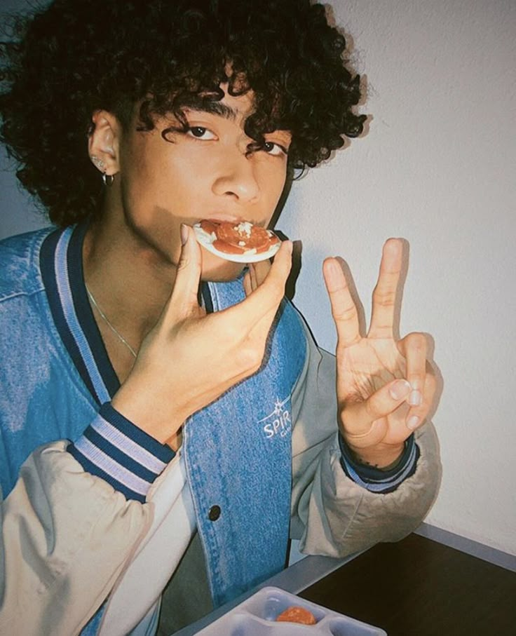 a young man with curly hair eating food and making the peace sign while sitting at a table