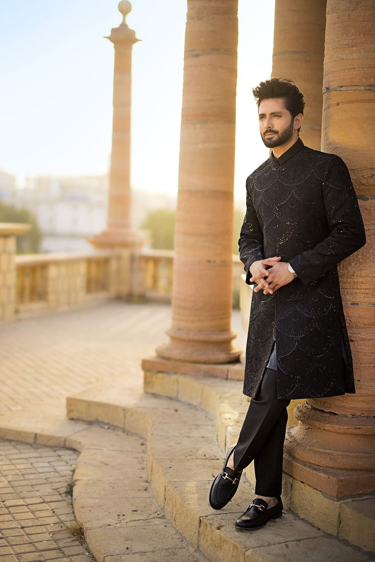 a man standing next to some pillars wearing a black suit and tie with his hands on his hips