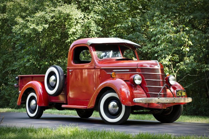 an old red truck is driving down the road in front of some trees and grass