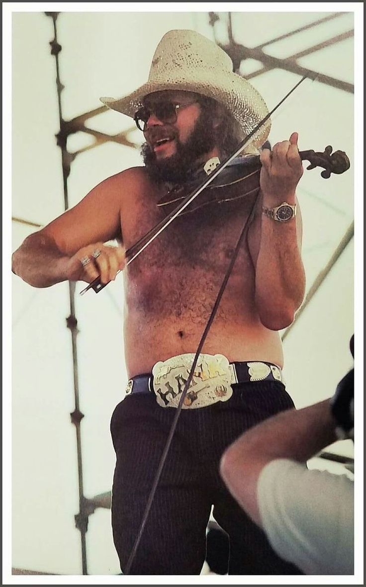 a man with no shirt on holding a violin in his right hand and wearing a cowboy hat