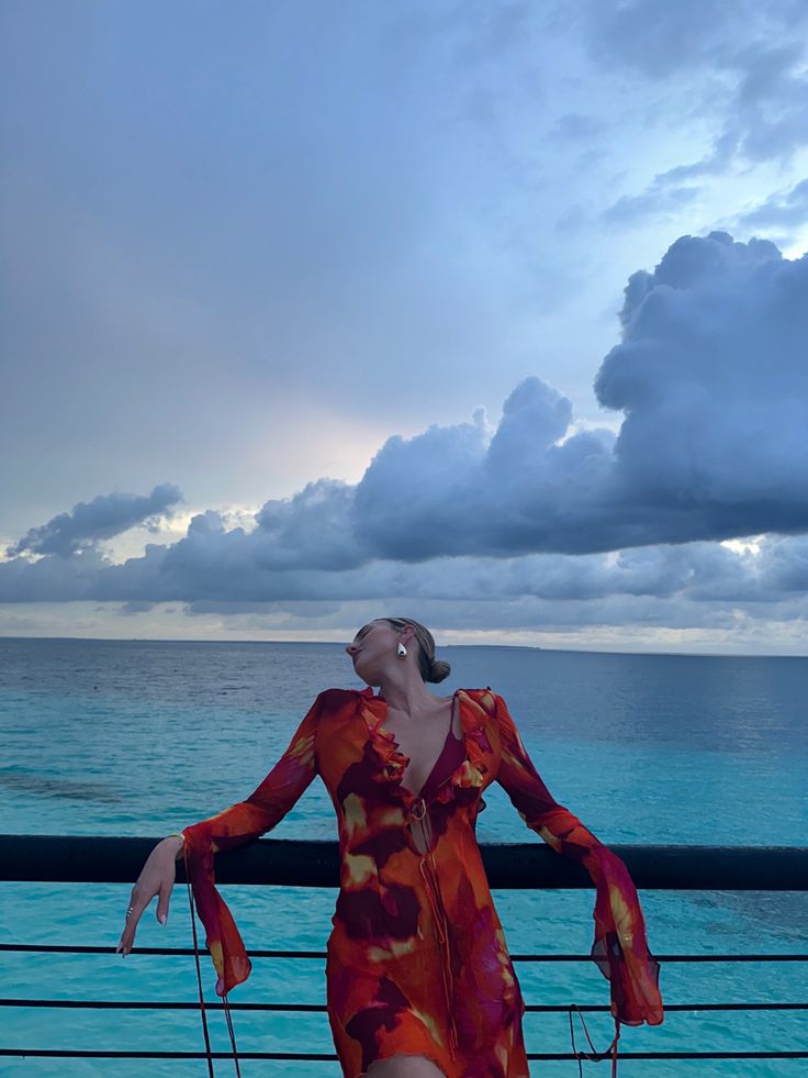 a woman in a red dress leaning on a railing looking up at the sky and water