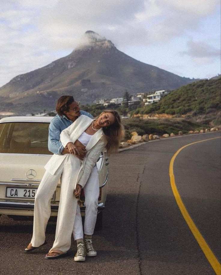 two women sitting on the back of a white car in front of a large mountain