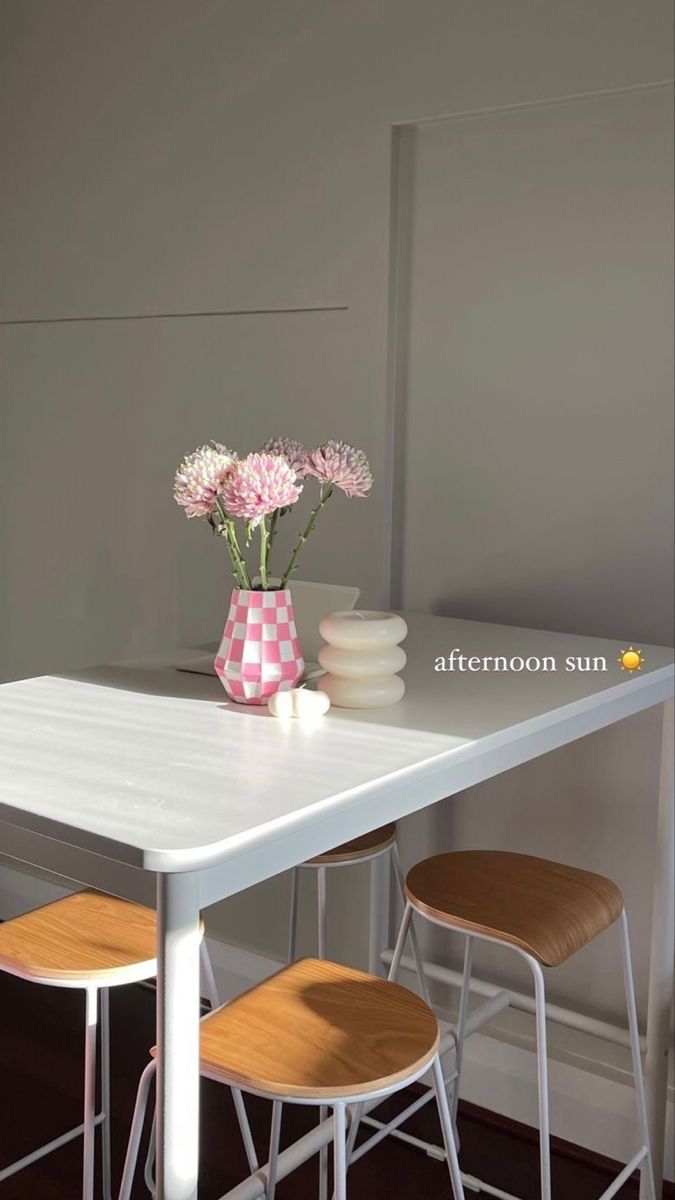 a white table with three stools and a pink vase on the top, in front of a gray wall