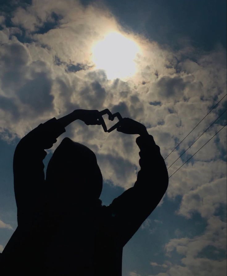 the silhouette of a person making a heart shape with their hands, against a cloudy sky