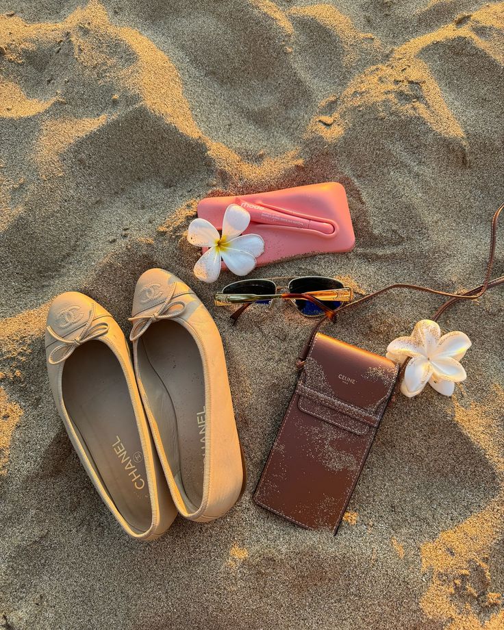 two pairs of shoes and sunglasses laying on the sand with flowers in front of them