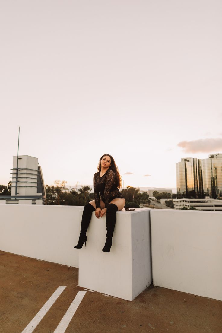 a woman sitting on top of a white wall next to a parking lot with tall buildings in the background