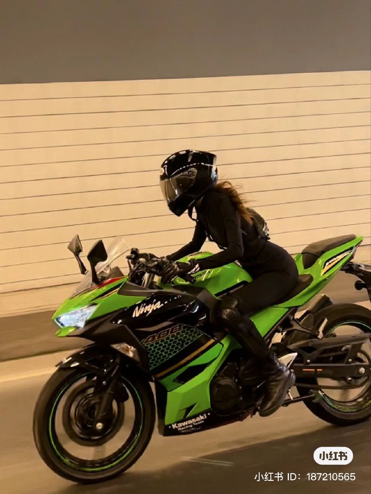 a woman riding on the back of a green motorcycle down a street in front of a white wall