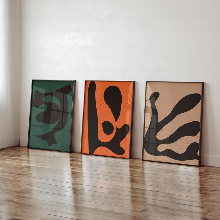 three framed art pieces sitting on top of a hard wood floor next to a window