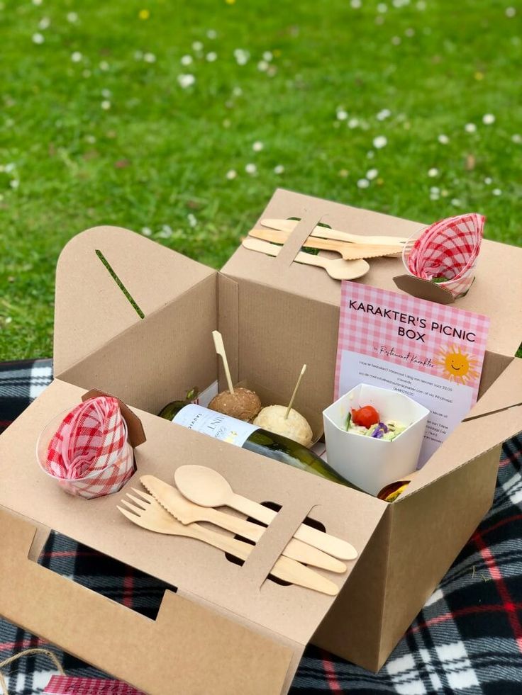 an open cardboard box with utensils and food in it on a plaid blanket