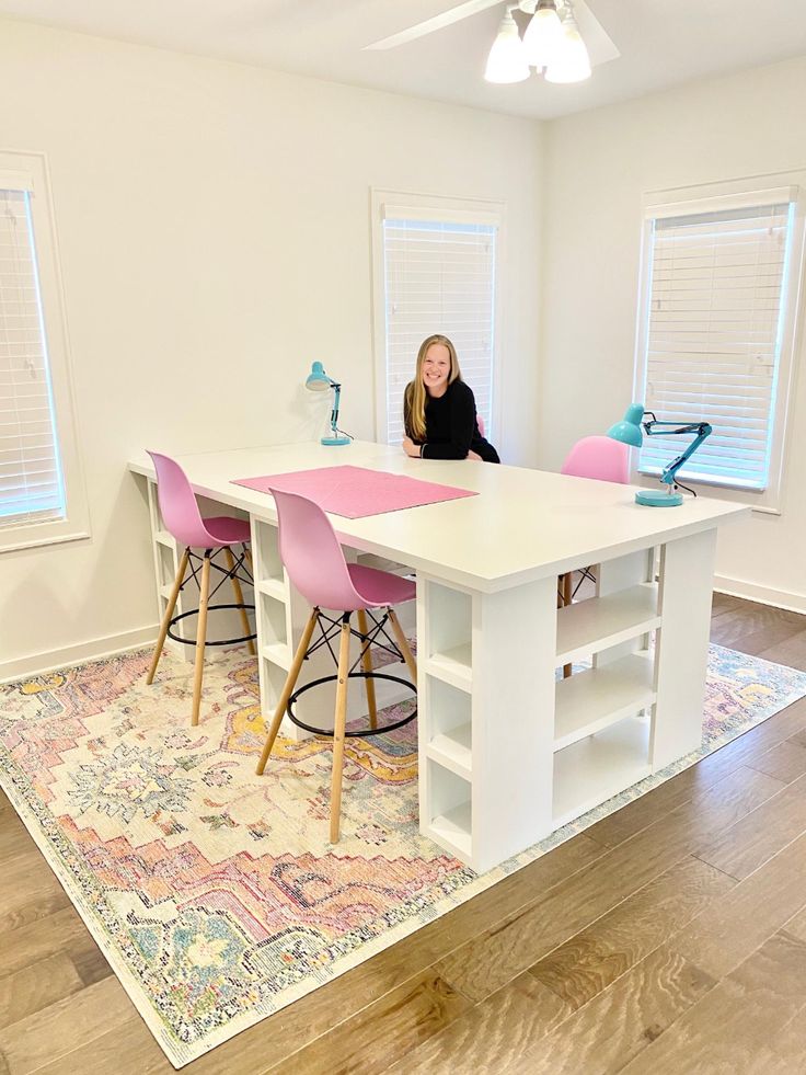 a woman sitting at a white table in a room with pink chairs and a rug on the floor