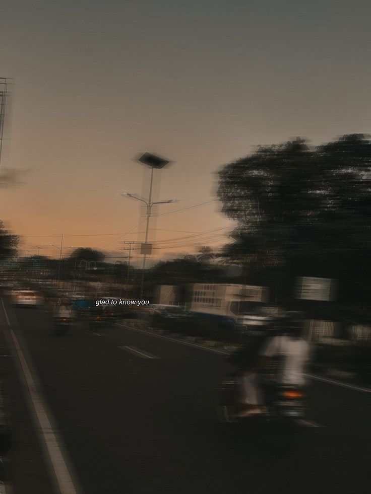 two people riding motorcycles down the street at dusk with buildings in the backgrouds