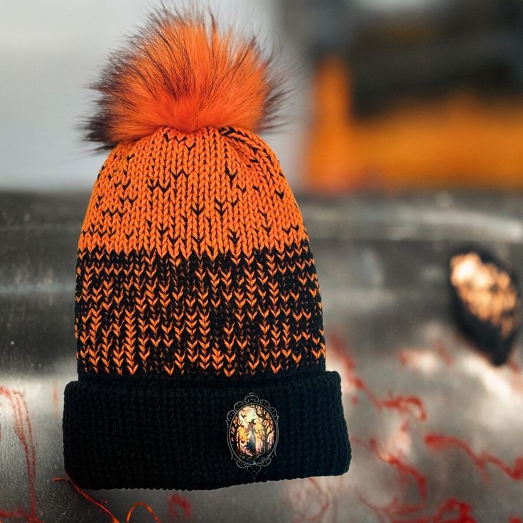 Handmade Knit Beanie with Large Orange Pom-Pom and Artistic Witch Gothic Badge - Black and Orange Winter Hat Stand out in any crowd with our handmade knit beanie, crafted for both warmth and style. This beanie features a striking black and orange pattern, perfect for making a bold fashion statement. Topped with a vibrant large orange faux fur pom-pom, this hat is both cozy and eye-catching. The gothic style artistic witch badge on the cuff adds a touch of elegance and creativity, making this beanie truly unique. Features: *Color: Black and orange pattern *Material: High-quality acrylic yarn for warmth and durability *Pom-Pom: Bright orange large faux fur pom-pom *Embellishment: Artistic badge on the cuff *Fit: One size fits most, with a comfortable and stretchy design *Care Instructions: H Black Knitted Hat For Halloween, Witchy Adjustable Winter Hats, Black Knitted Beanie In Acrylic Yarn, Black Acrylic Yarn Beanie Cap, Black Acrylic Yarn Beanie, Halloween Beanie One Size Fits Most, Black Yarn Beanie, Black Knitted Bonnet, One Size Fits Most, Black Knitted Bonnet One Size