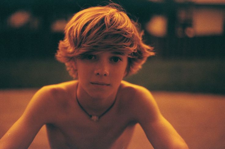 a shirtless boy sitting on the ground with his hands behind his head and looking at the camera