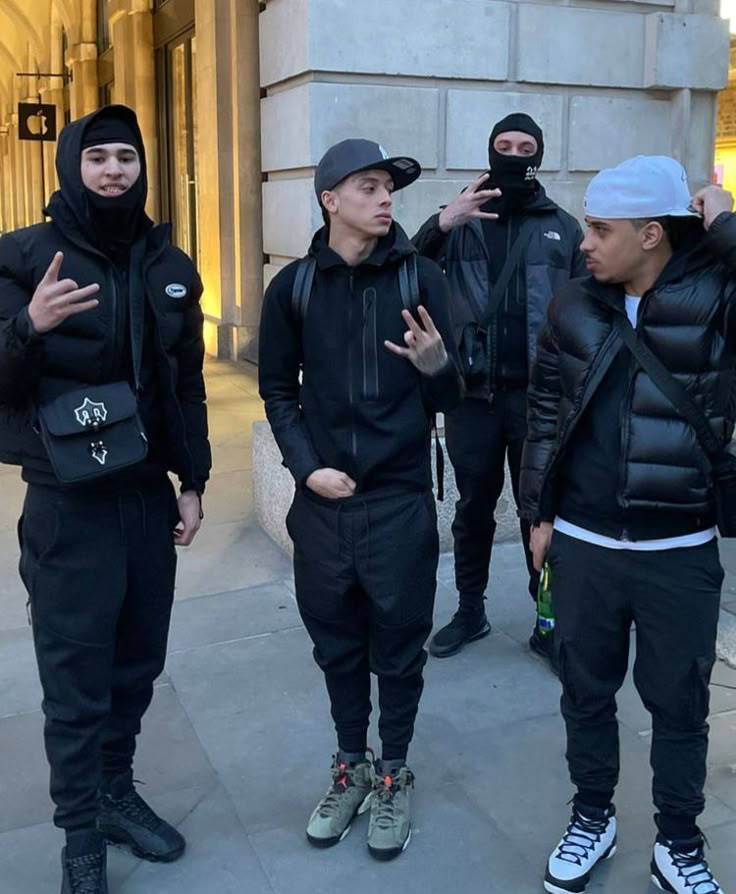 four young men in black hoodies standing on the sidewalk with their hands up and looking at something