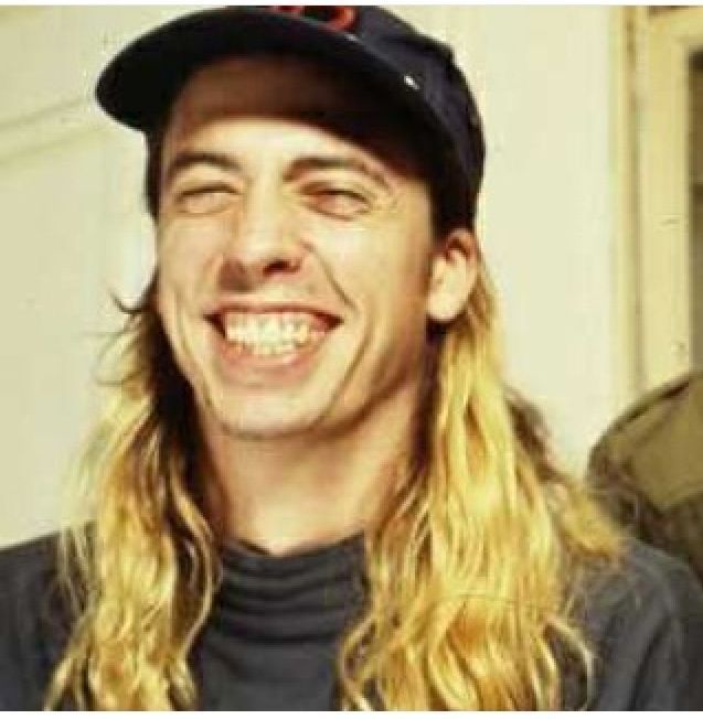 a man with long blonde hair wearing a baseball cap and black shirt smiling at the camera