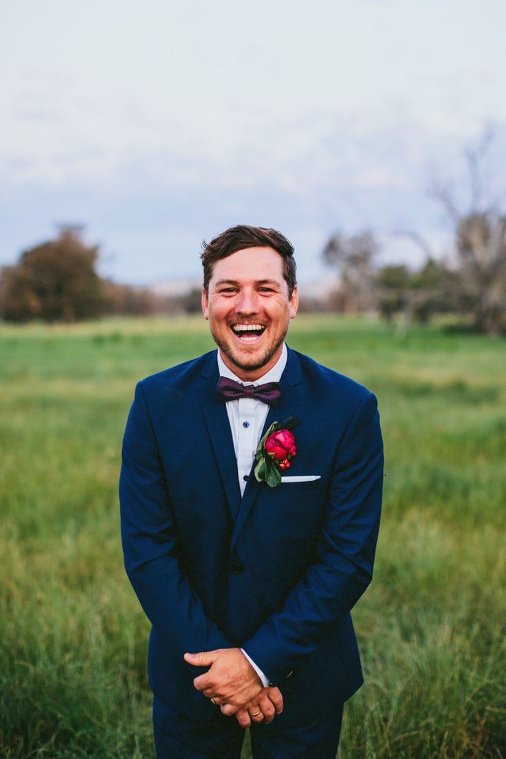 a man in a tuxedo smiles as he stands in a field with his hands on his hips