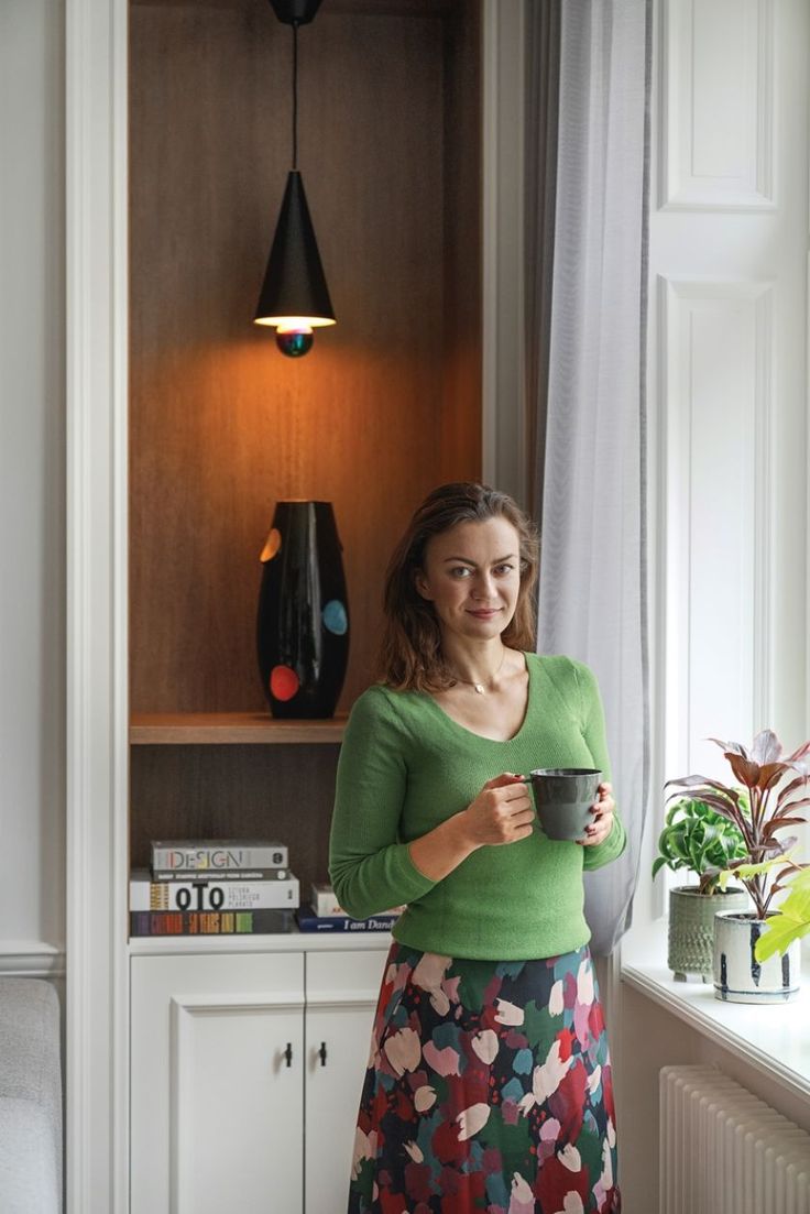 a woman standing in front of a window holding a cup and looking at the camera