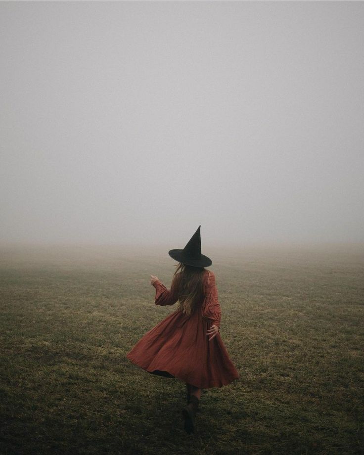 a woman in a red dress and black hat is walking through the foggy field