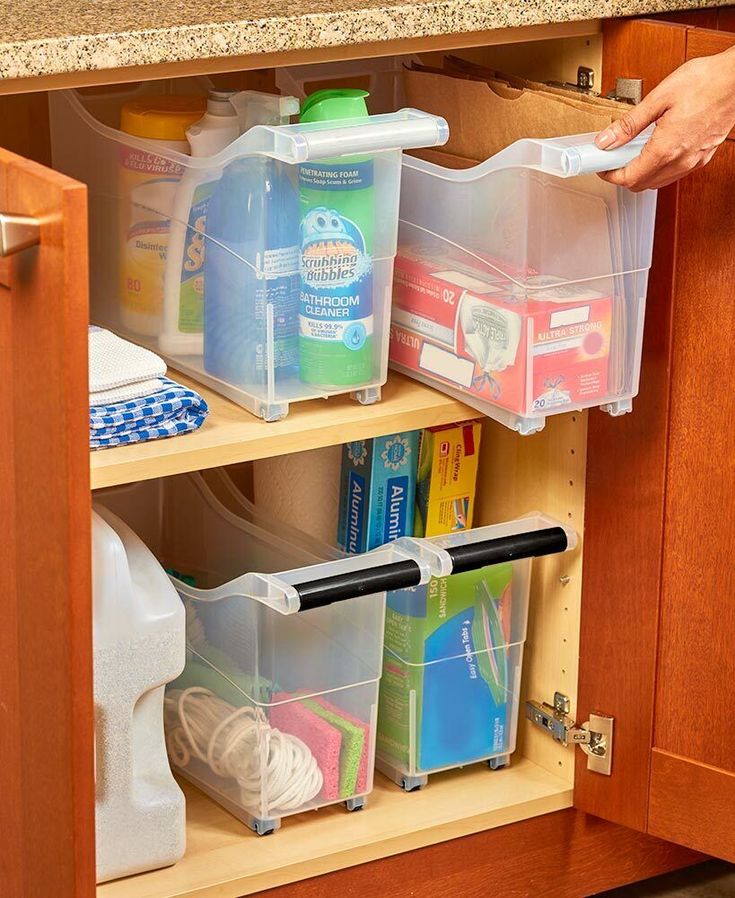 a kitchen cabinet with clear plastic bins holding cleaning supplies