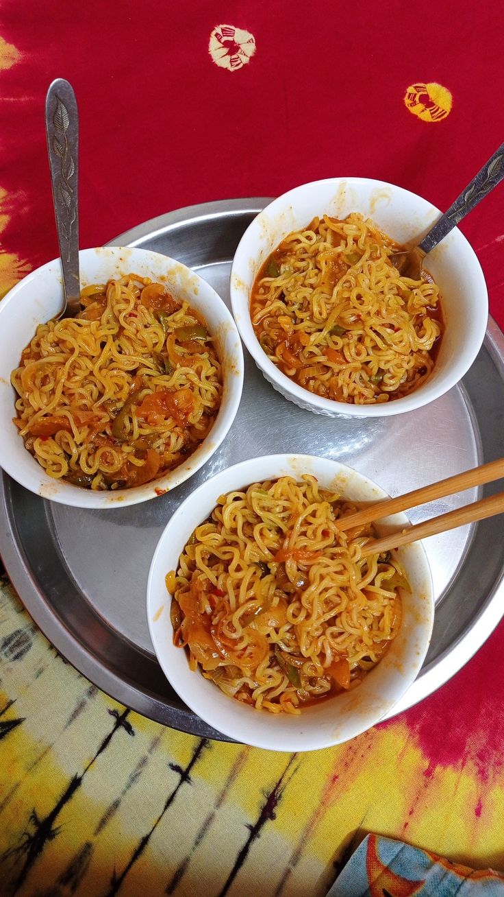 three bowls of noodles with chopsticks in them on a metal tray and red table cloth